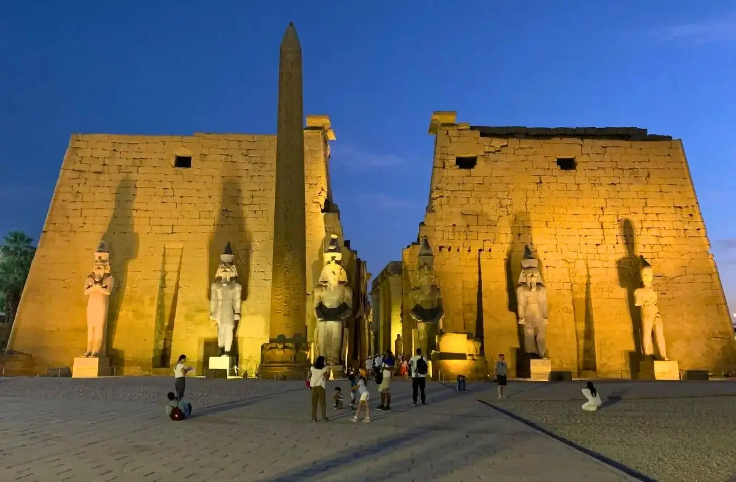 The illuminated entrance of the Temple of Luxor in Egypt at night, featuring a tall stone obelisk and colossal statues of Ramesses II guarding a massive pylon gateway under a deep blue twilight sky.