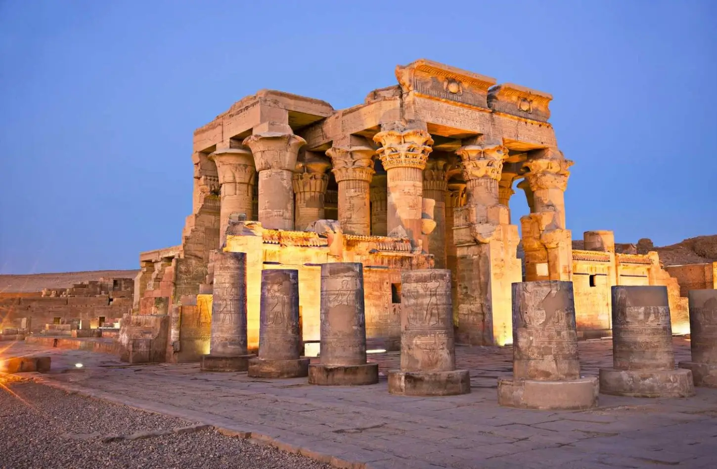A wide shot of the Kom Ombo temple at dusk, featuring glowing stone columns with ancient Egyptian carvings under a deep blue evening sky