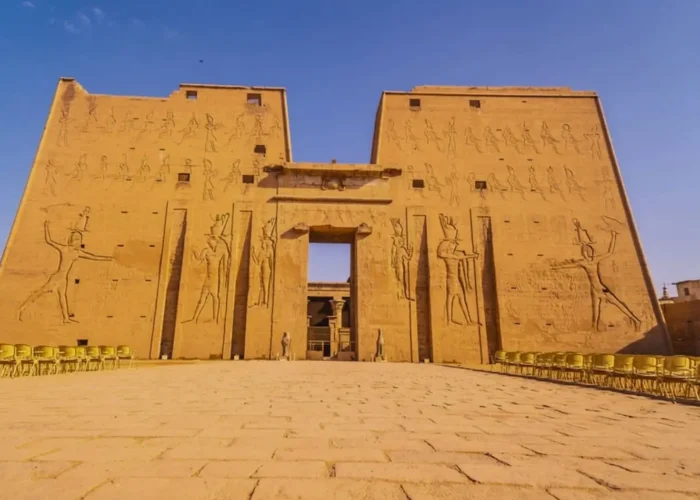 Ancient stone ruins and detailed hieroglyphic carvings in the foreground of the Edfu Temple Egypt, looking toward the main monumental gateway and a surviving ceremonial column.