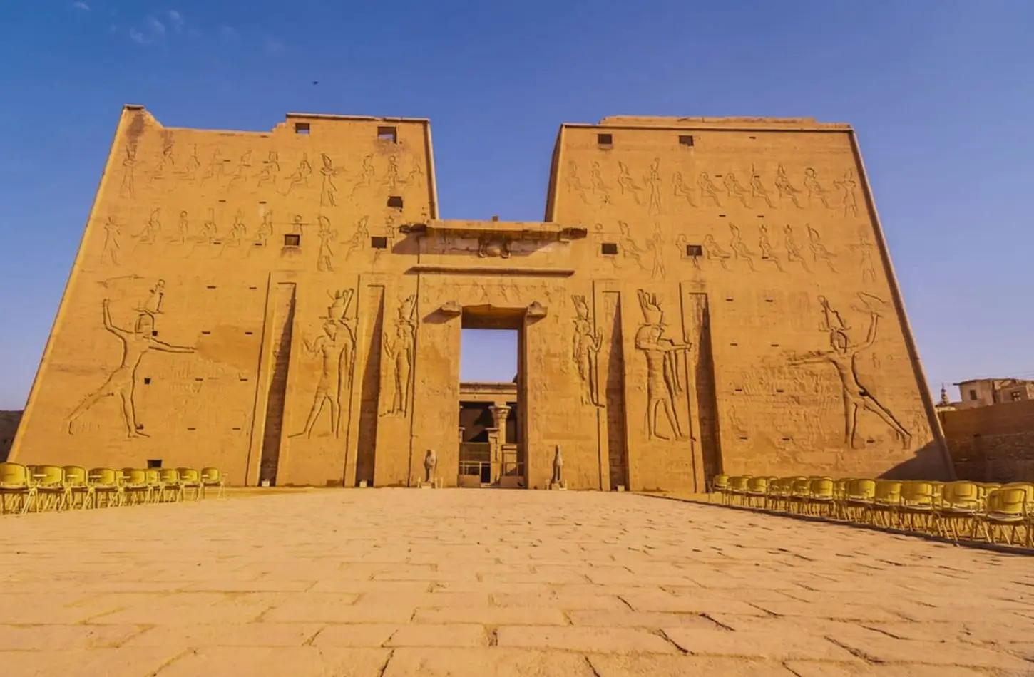 Ancient stone ruins and detailed hieroglyphic carvings in the foreground of the Edfu Temple Egypt, looking toward the main monumental gateway and a surviving ceremonial column.