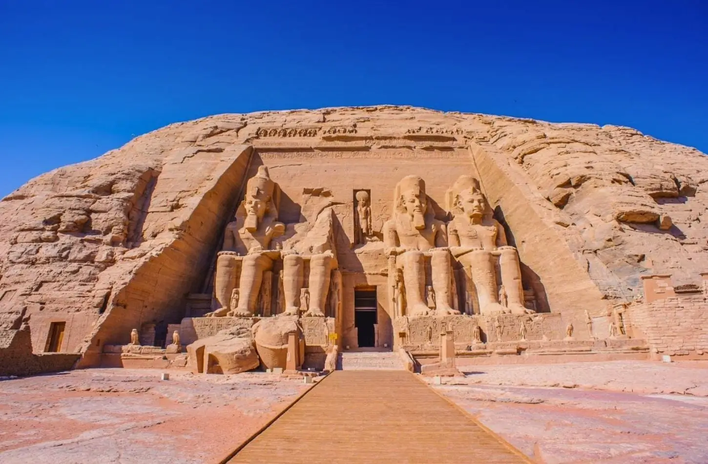 A wide-angle view of the Great Abu Simbel Temple in Egypt, featuring four colossal seated statues of Ramesses II carved into a sandstone cliff under a clear blue sky.